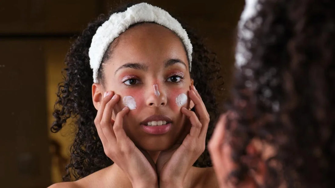 Woman applying cream to her face with a towel on her head, looking into a mirror.