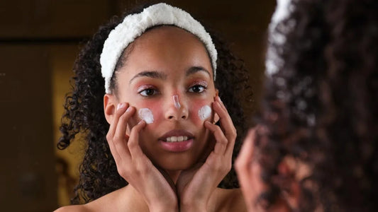 Woman applying cream to her face with a towel on her head, looking into a mirror.